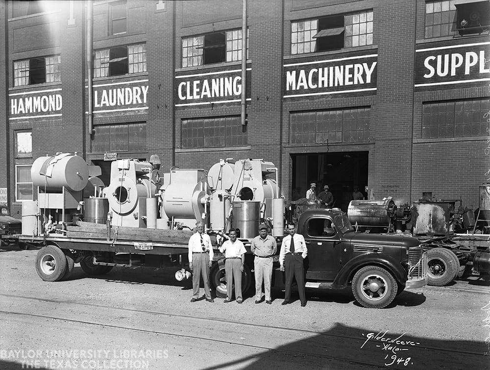 Men stand by a truck loaded with industrial machinery outside a building labeled
