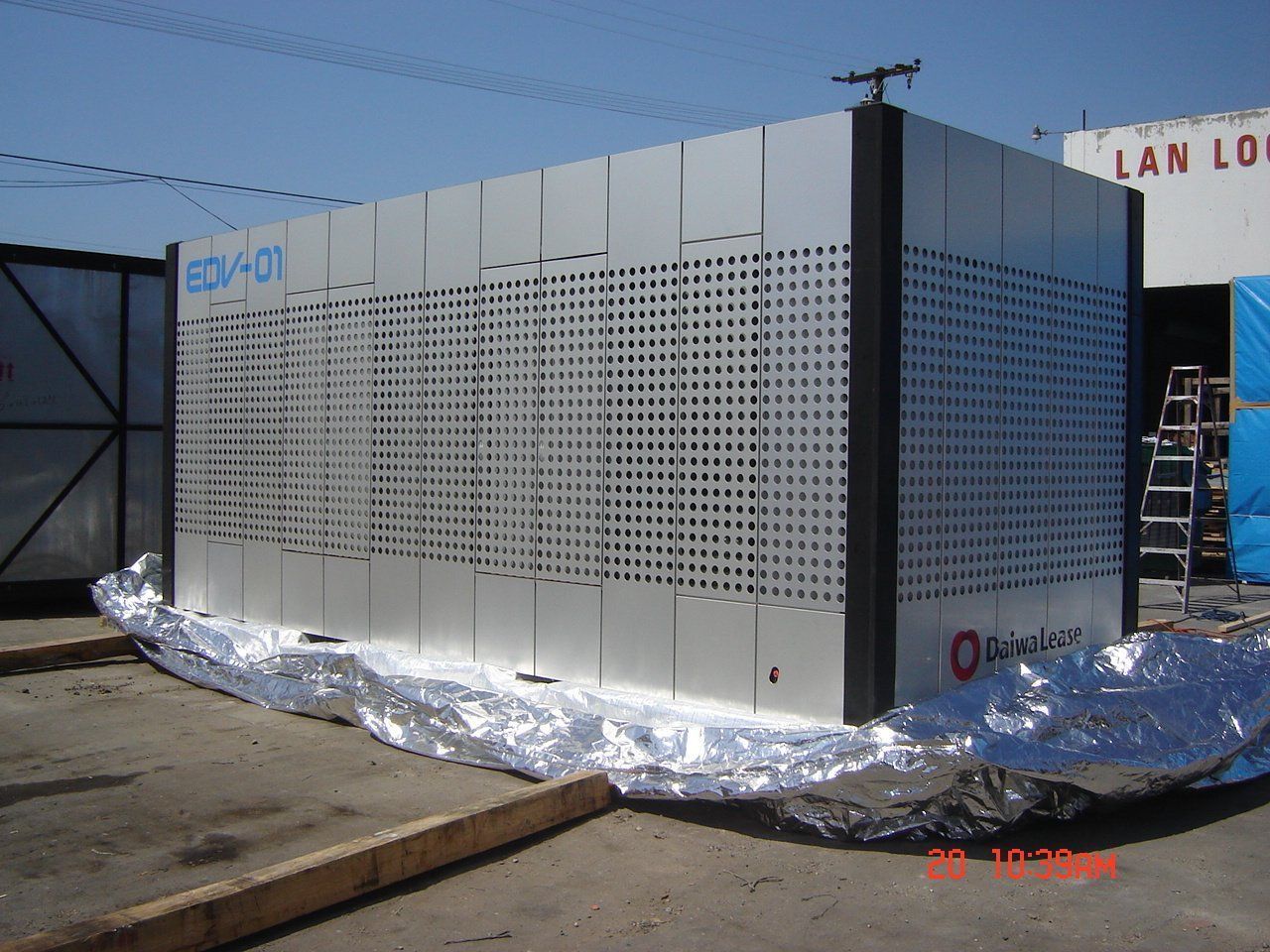 Large, rectangular industrial unit with perforated panels, likely a machine, sitting outdoors on reflective material.