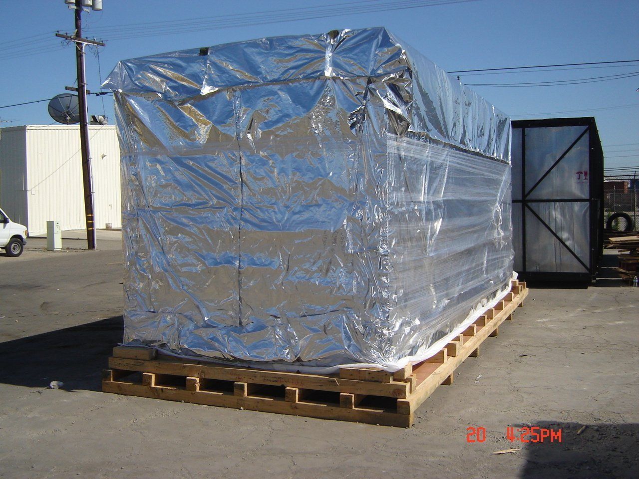 Large rectangular package wrapped in reflective silver material on a wooden pallet outdoors.