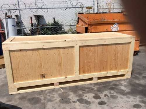 Wooden shipping crate on a concrete surface, near an orange dumpster and chain-link fence.