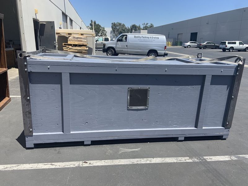 Gray wooden crate with a small window, parked on pavement. A white van is in the background.