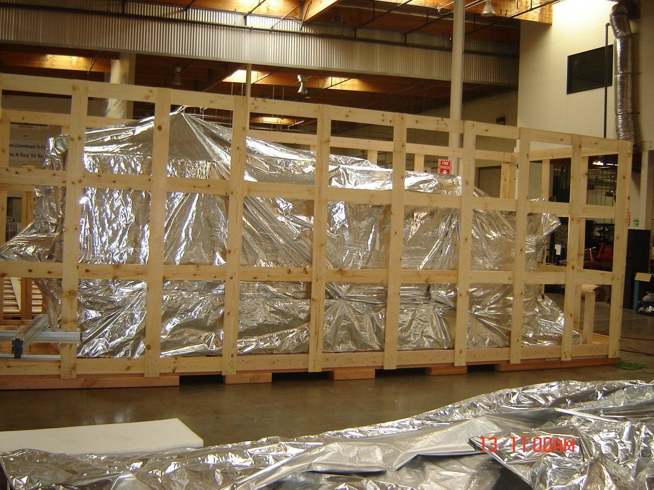Large object wrapped in silver foil inside wooden shipping crate, in a warehouse.