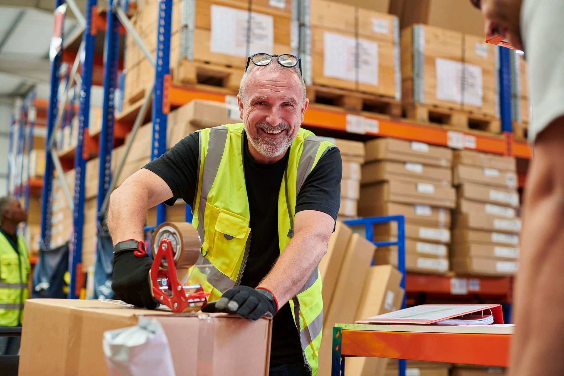 Warehouse worker tapes a box, smiling. He wears a reflective vest and gloves; shelves of boxes in the background.