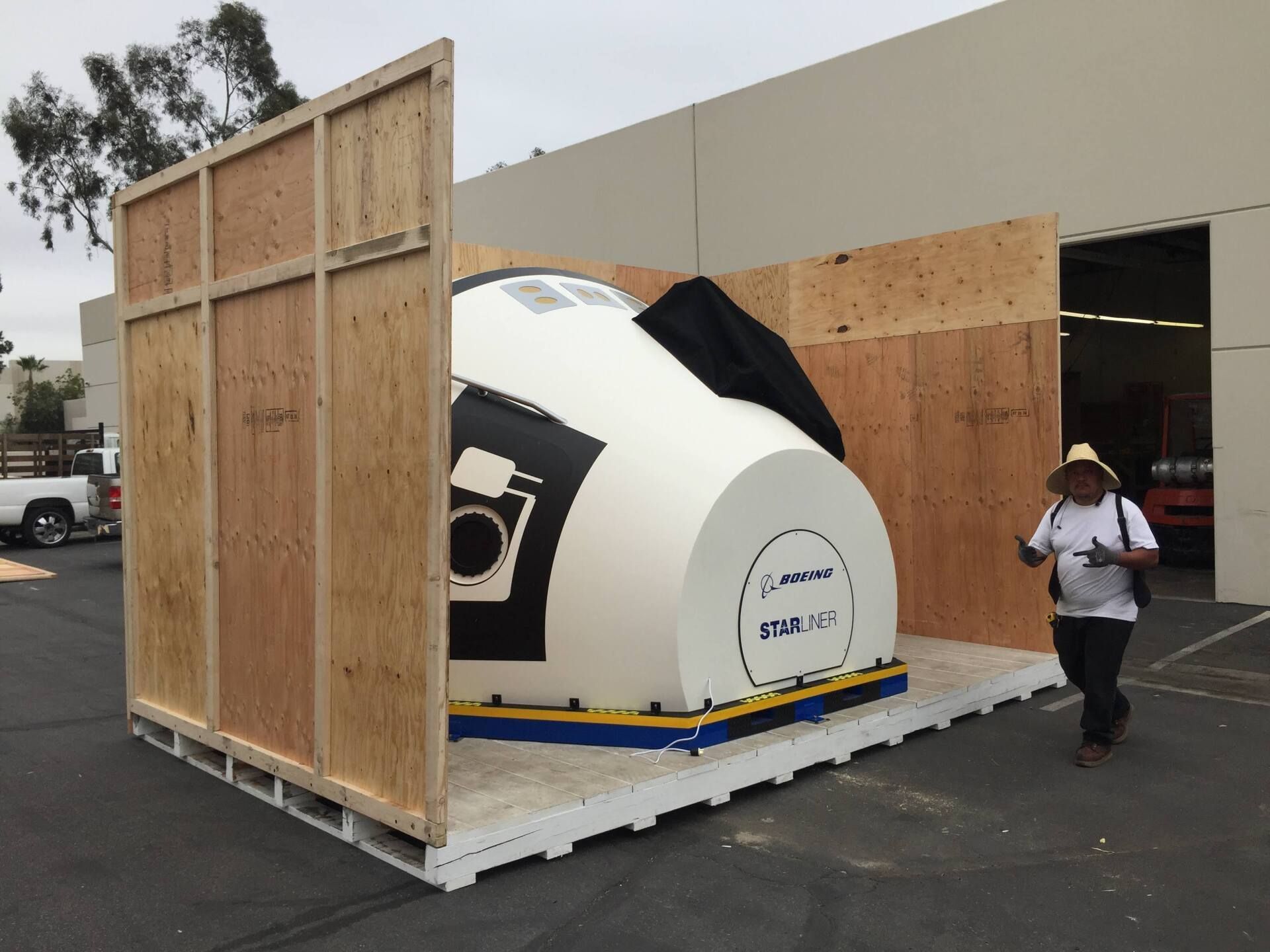 A white and black simulator is in a wooden crate, with a person walking beside it. Outdoor setting.