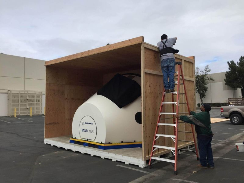 Two men packing a large white machine into a wooden crate outdoors. Cloudy sky overhead.