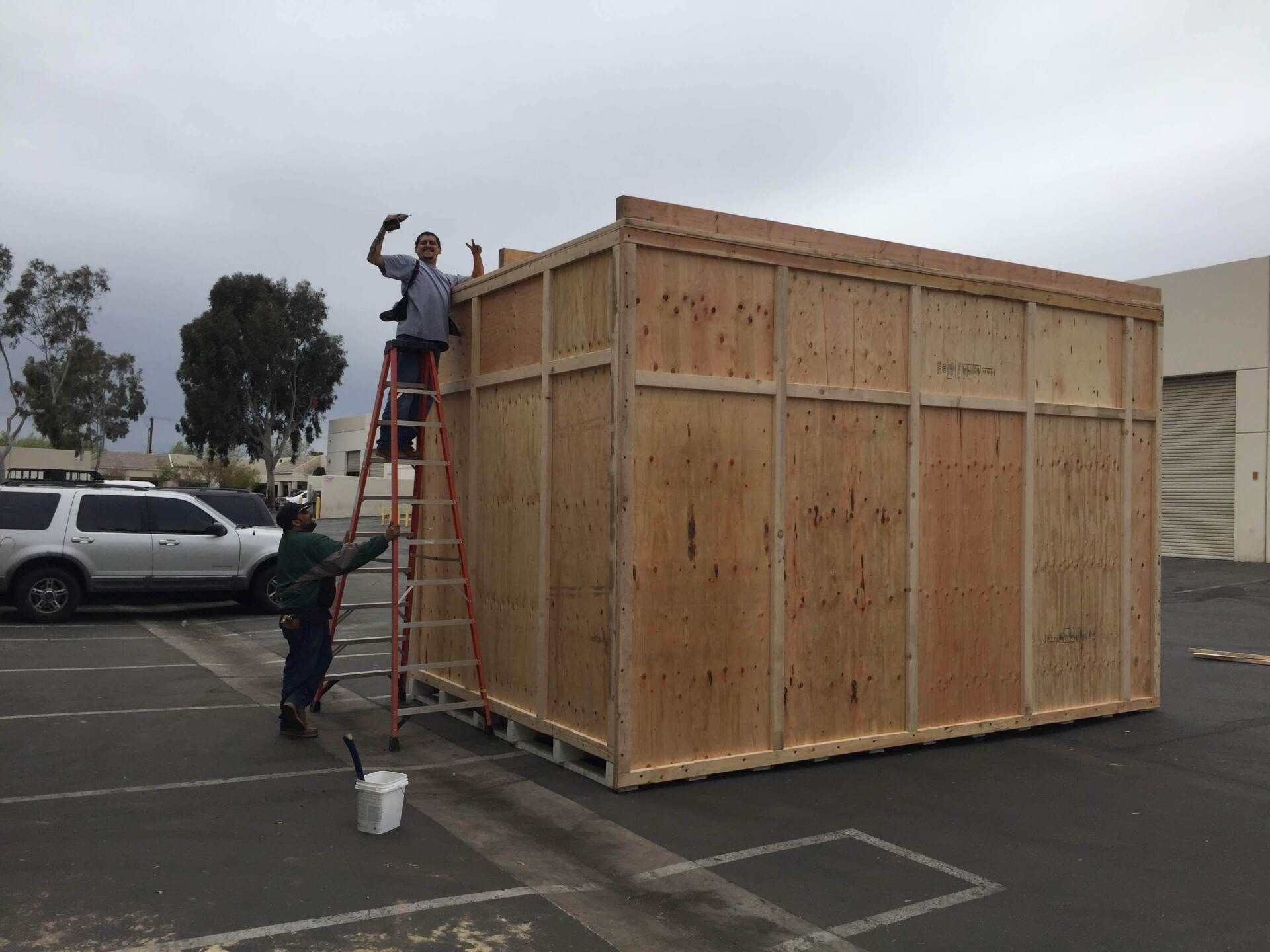 Two people constructing a large wooden crate outdoors; one on a ladder, one at the base.