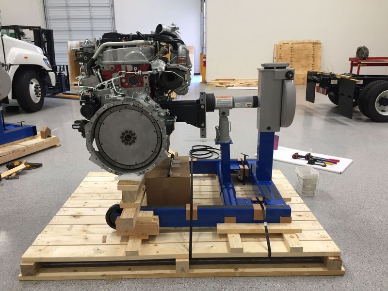 Engine mounted on a blue stand with tools, sitting on wooden pallets in a warehouse.