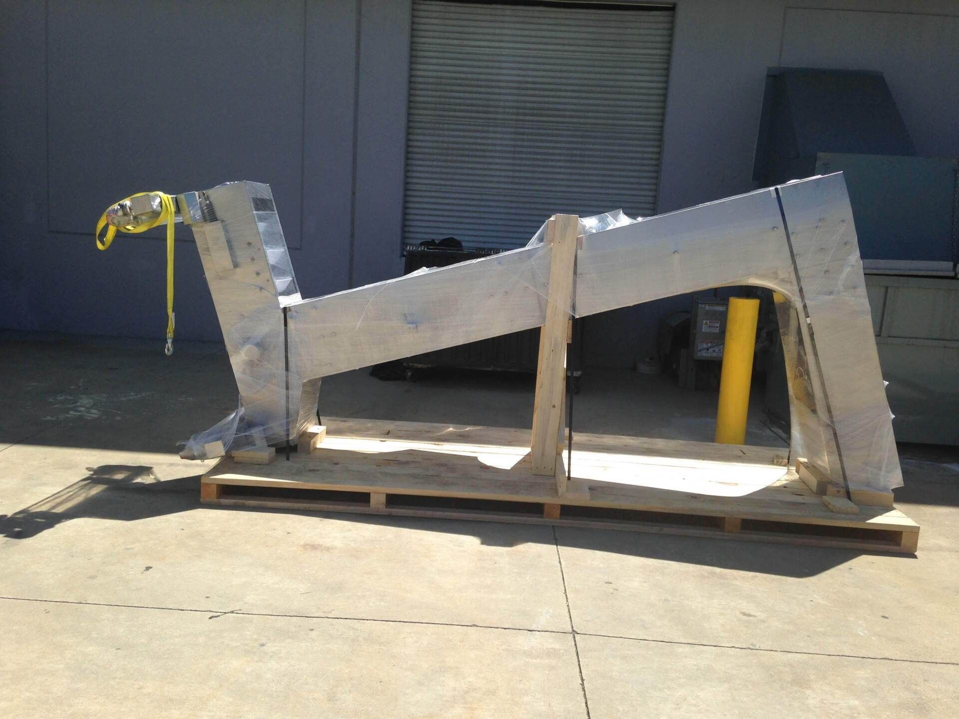 Large silver metal structure on a wooden pallet, outdoors. Yellow strap securing the structure.