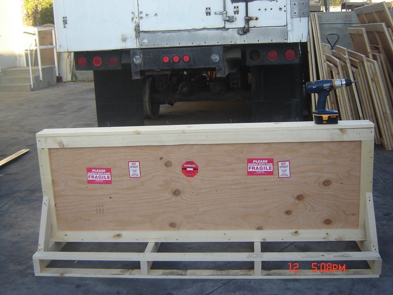 Wooden crate with plywood front, in front of a truck. A drill is on top of the crate.