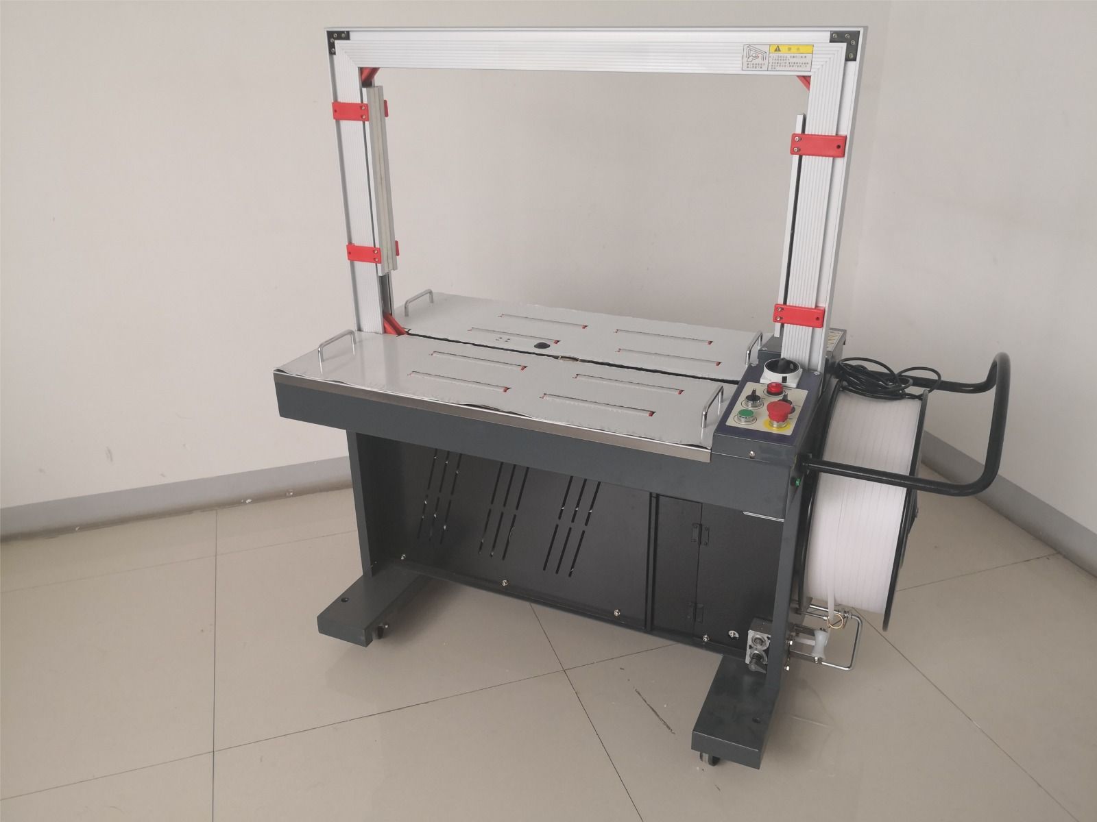 Flejadora automática, metal gris con mesa blanca, detalles en rojo, panel de control, rollo de flejado de plástico blanco.