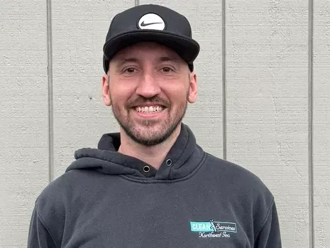 Man smiling, wearing a black cap with a Nike logo, black hoodie, in front of a gray wall.