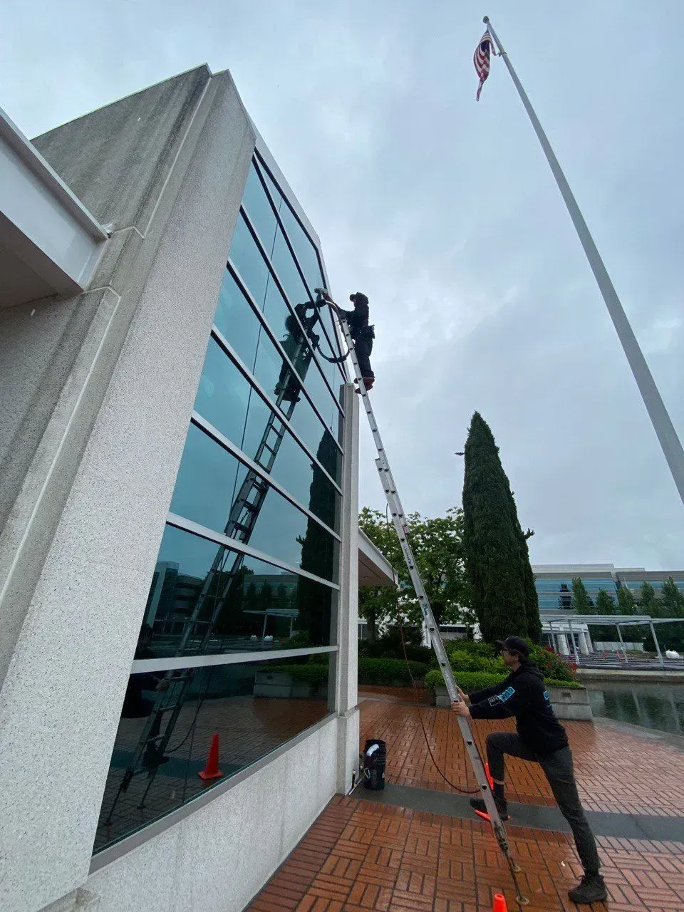 Man steadies ladder as window washers clean exterior windows of a building.
