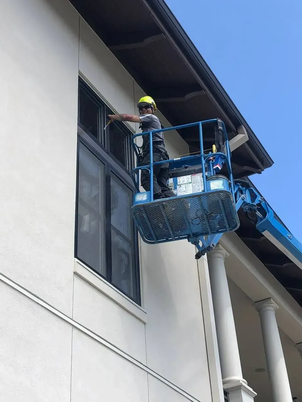 Worker in a lift, cleaning exterior window of a white building with dark window frames, on a sunny day.