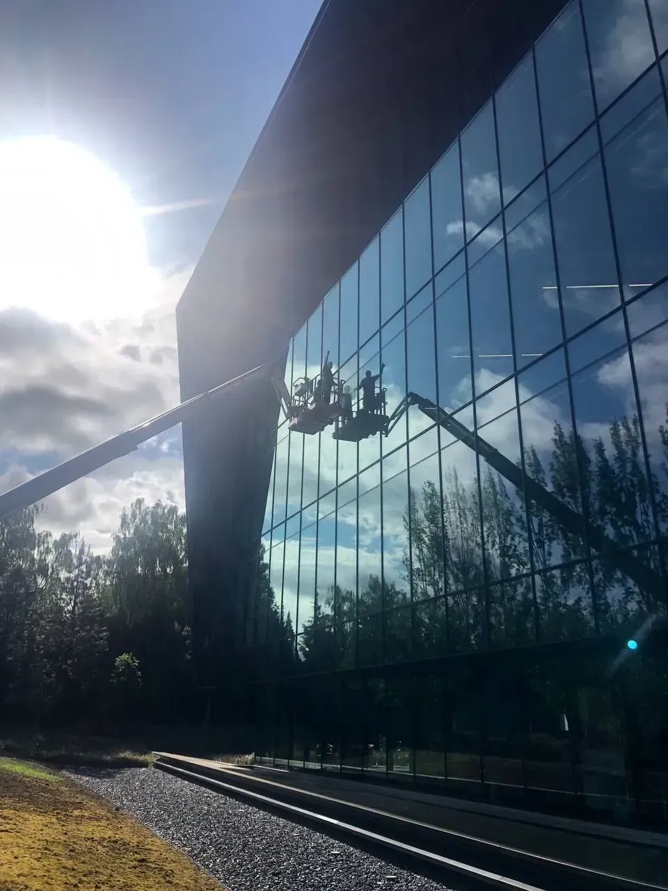 Workers in cherry pickers cleaning large glass windows of a modern building; sunlight reflects.