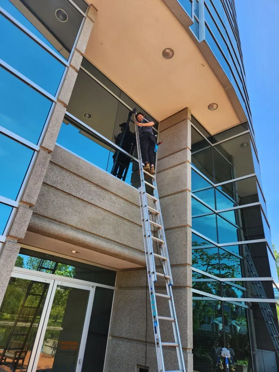 Person on a tall ladder cleaning windows of a modern building with reflective glass. Sunny day.