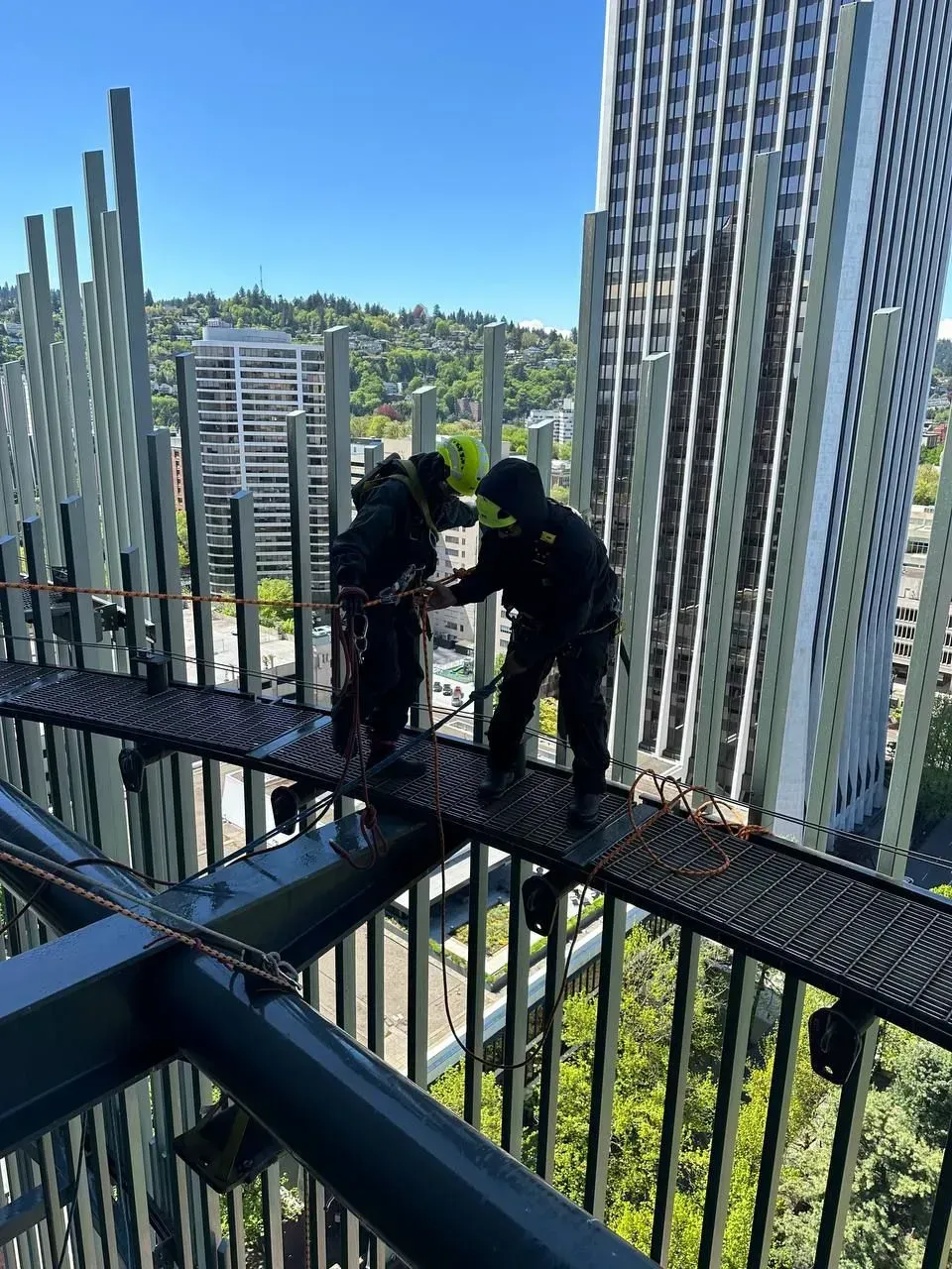 Two workers on a high platform, inspecting a structure. One is adjusting the other's gear with tall buildings and trees in the background.