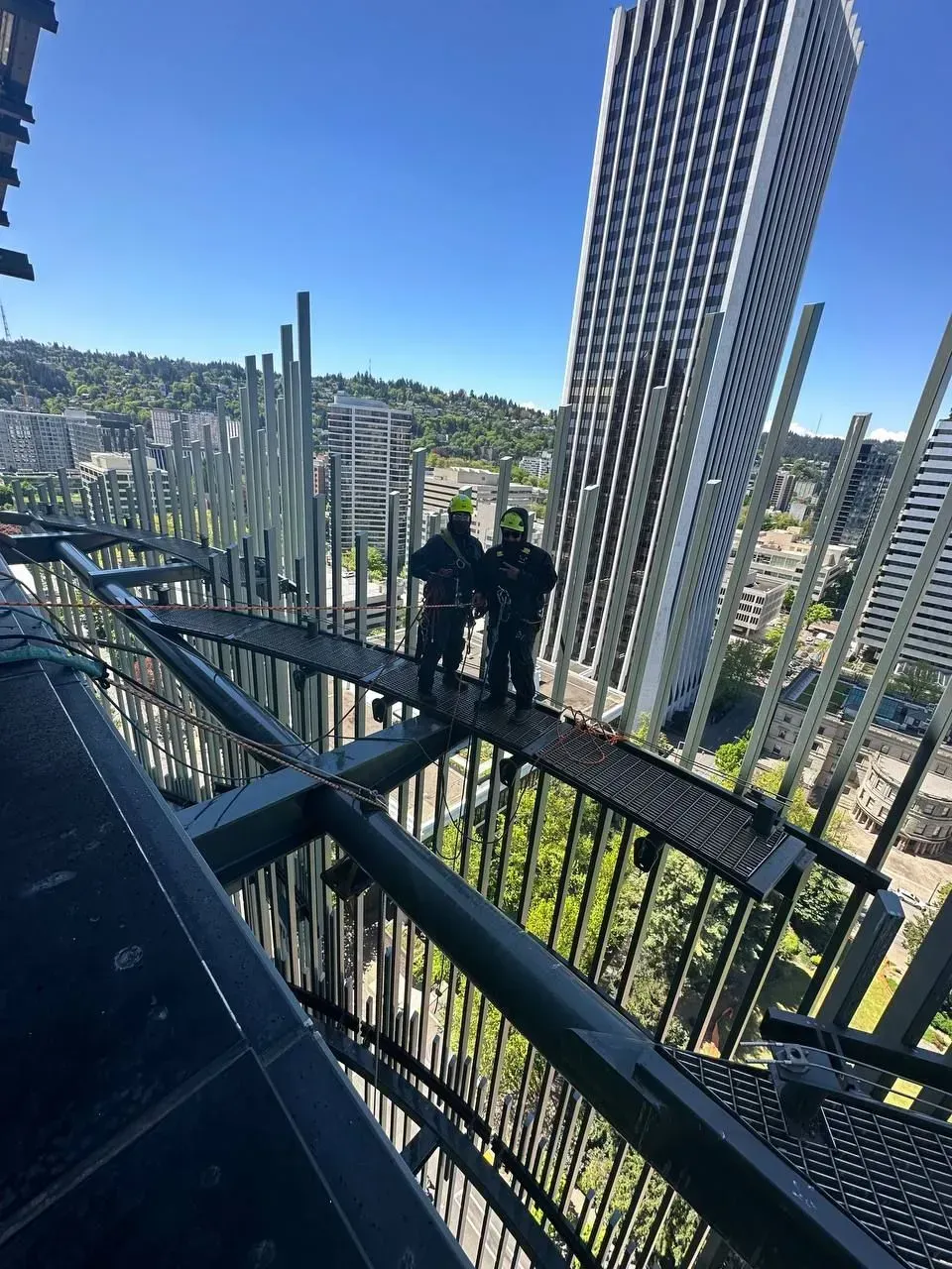 Two people stand on a narrow bridge high above buildings. They wear dark uniforms. Sunny day.