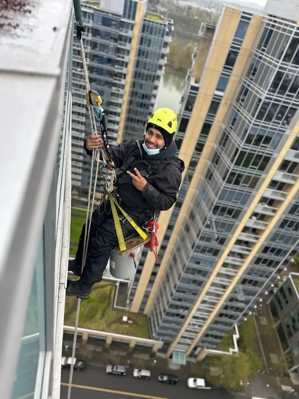 Window washer suspended from a high-rise building, smiling, wearing a helmet and safety gear, with urban background.