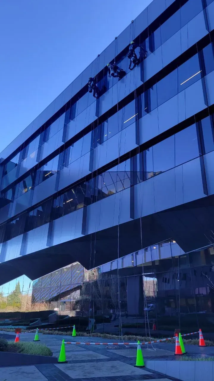 Window washers on a dark blue glass building under a clear sky. Traffic cones line the ground below.
