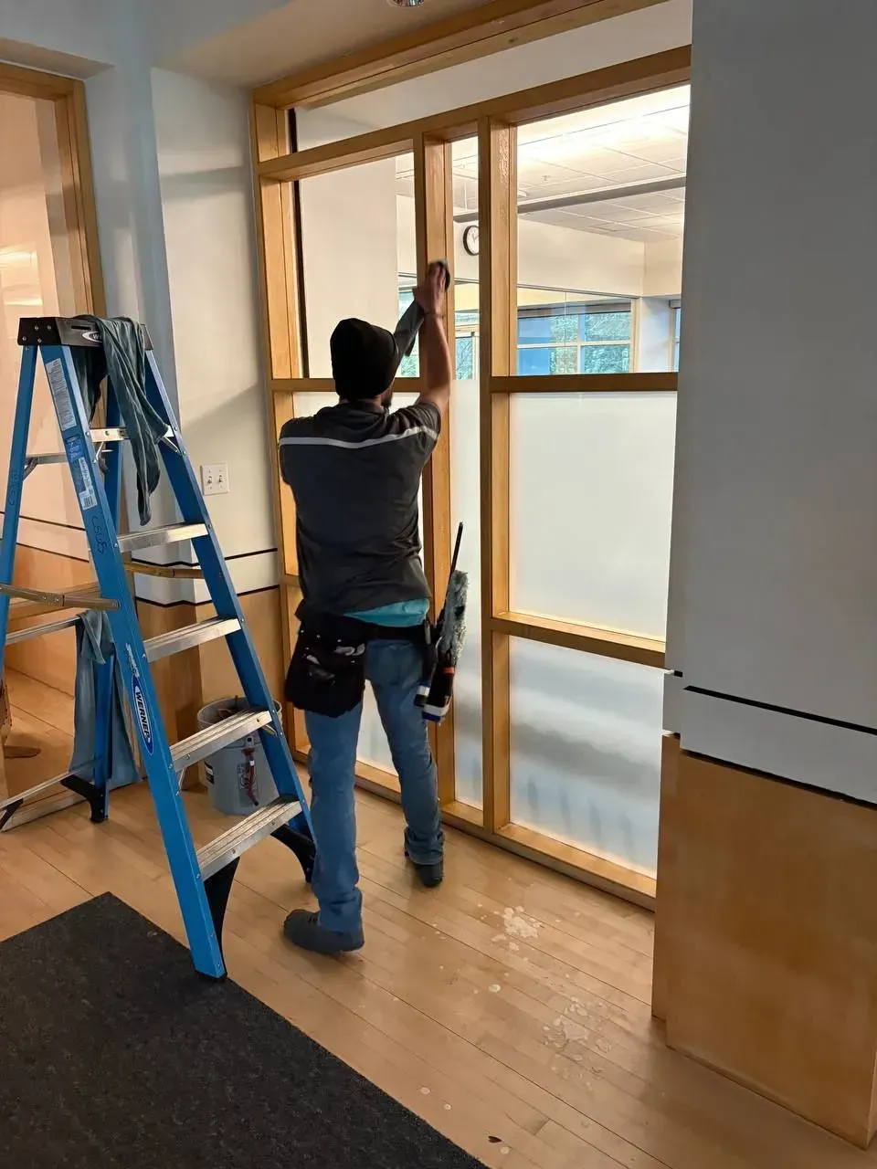 Person installing window film, standing by a blue ladder. Frosted glass panels are framed in light wood.