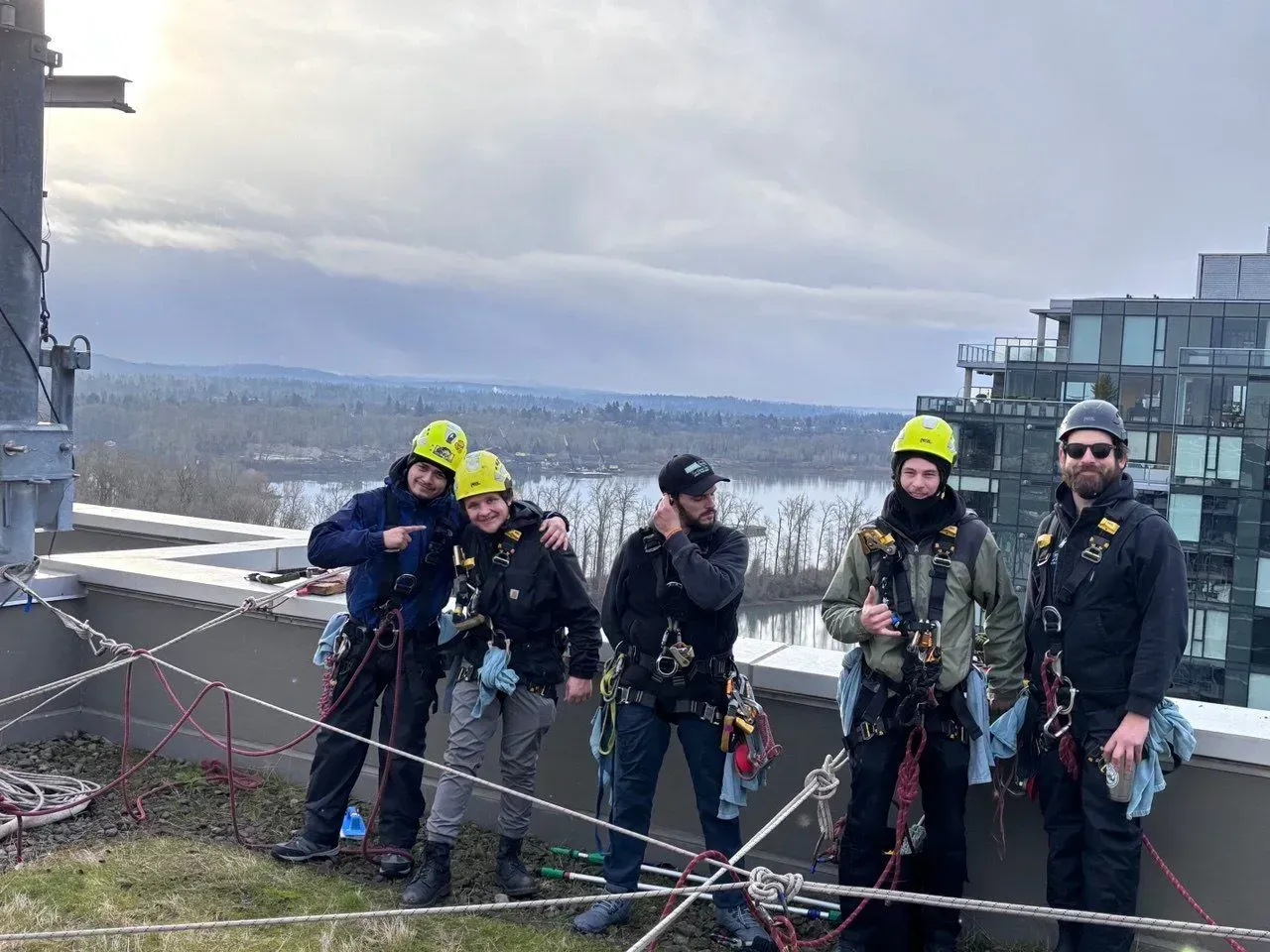 Five people in safety gear pose on a rooftop with a lake and city in the background.