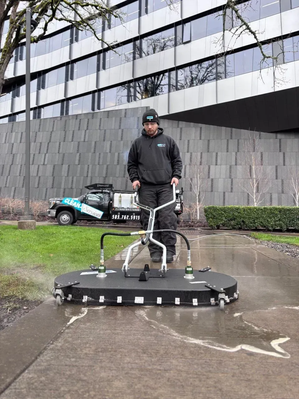 Man using a surface cleaner on a sidewalk in front of a building. A truck is parked nearby.