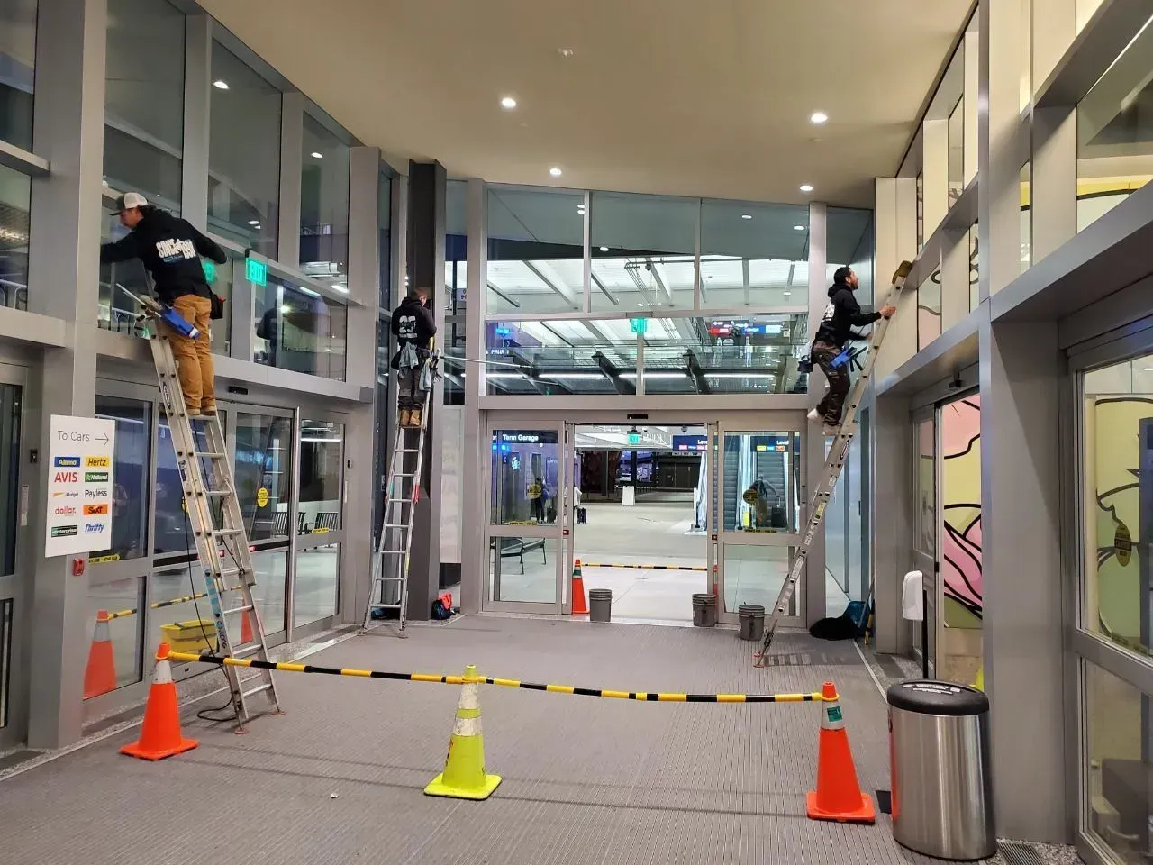 Three workers on ladders cleaning windows inside an airport terminal entrance.