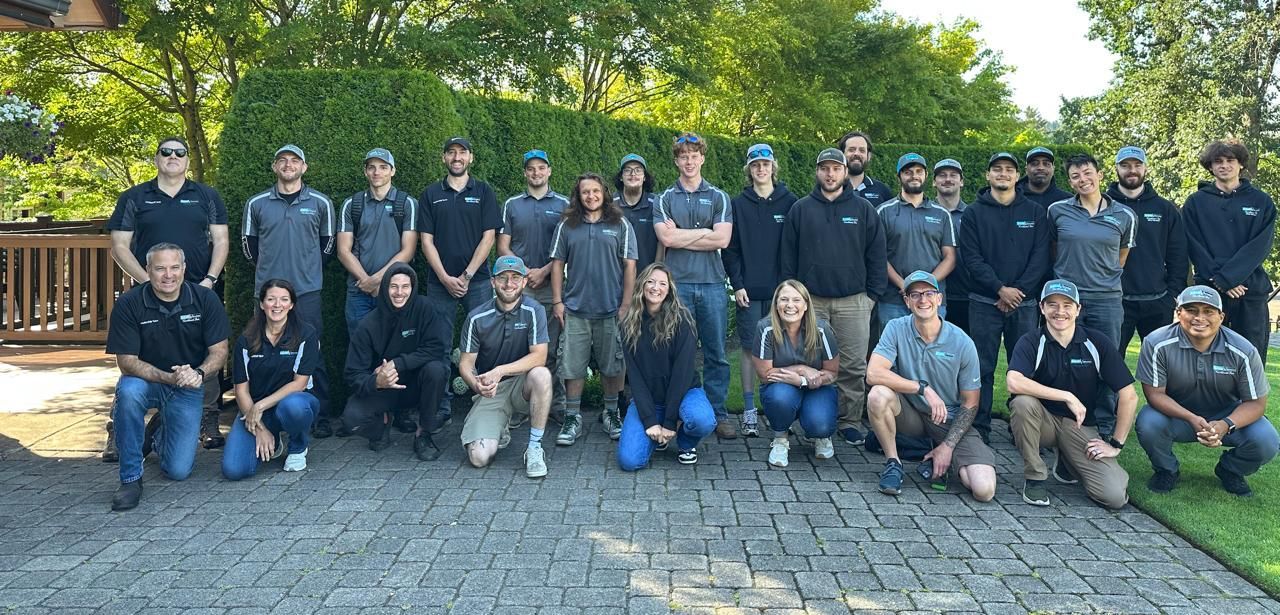 Group of people in casual attire posing outdoors. Brick paved area, trees in the background.
