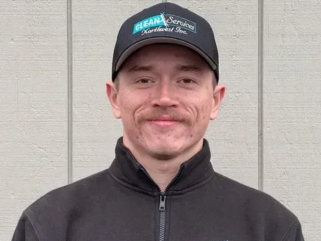 Man in black cap and jacket, smiling, standing in front of a light-colored building.
