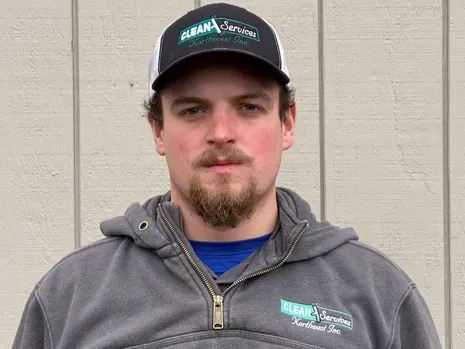 Man wearing a baseball cap and gray hoodie, looking at the camera. He has a beard and is standing outside.