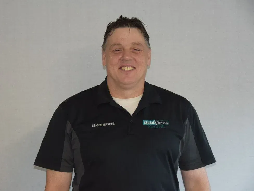 Man in black polo shirt smiles in front of a gray wall.