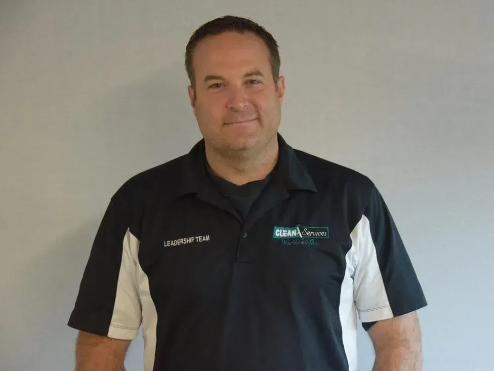 Man in black polo shirt with logo, standing against a gray wall, smiling slightly.