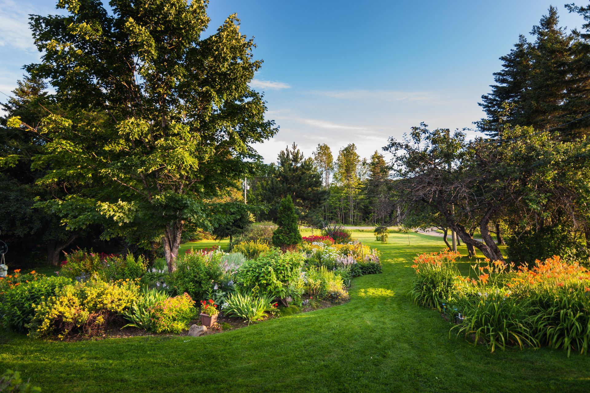 Lush green garden with flowerbeds, trees, and path leading towards sunlit background under blue sky.