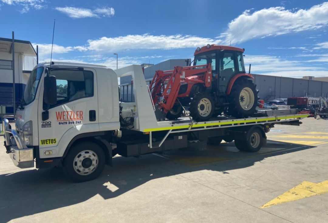 Tow Truck With A Red Tractor On The Back Is Parked In A Parking Lot — Wetzler Pty Ltd In Marmor QLD