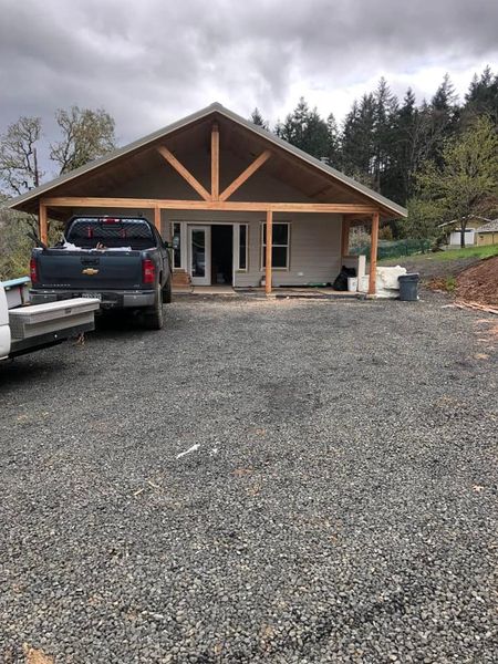 A light-colored building under construction with a wooden porch, gray gravel driveway, and a pickup truck.