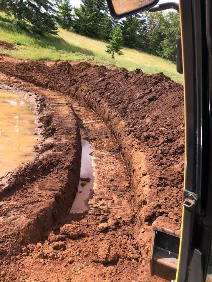Trench dug in muddy ground by a yellow construction vehicle, near a water-filled area and a grassy hill.