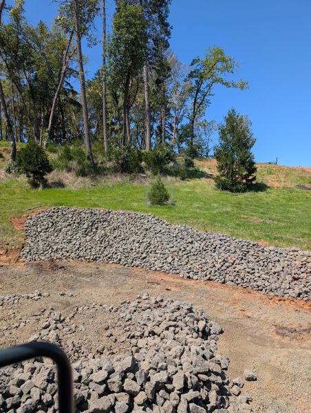 Stone retaining wall on a grassy hillside, with trees in the background under a blue sky.