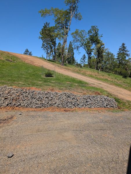 Stone retaining wall with gravel and grass leading up to a dirt path and trees under a blue sky.