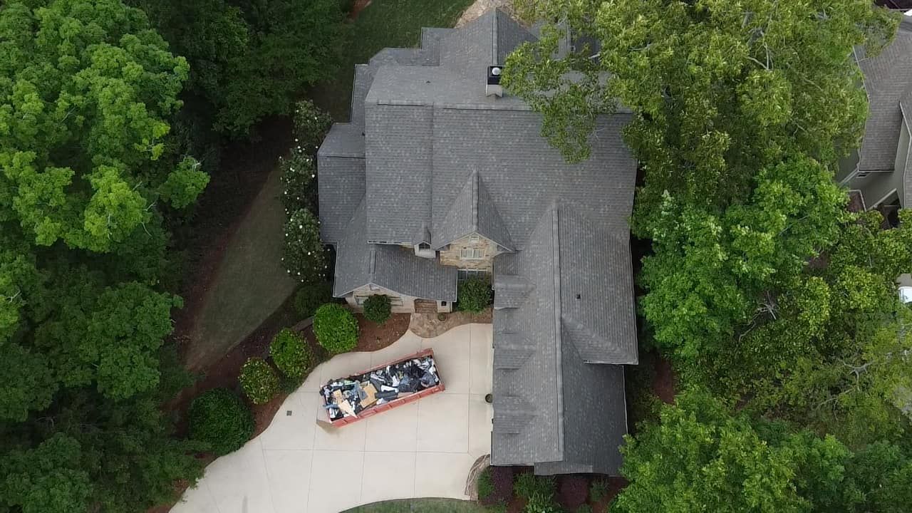 Aerial view of a gray-roofed house beside trees, with a white driveway and parked car near the entrance.