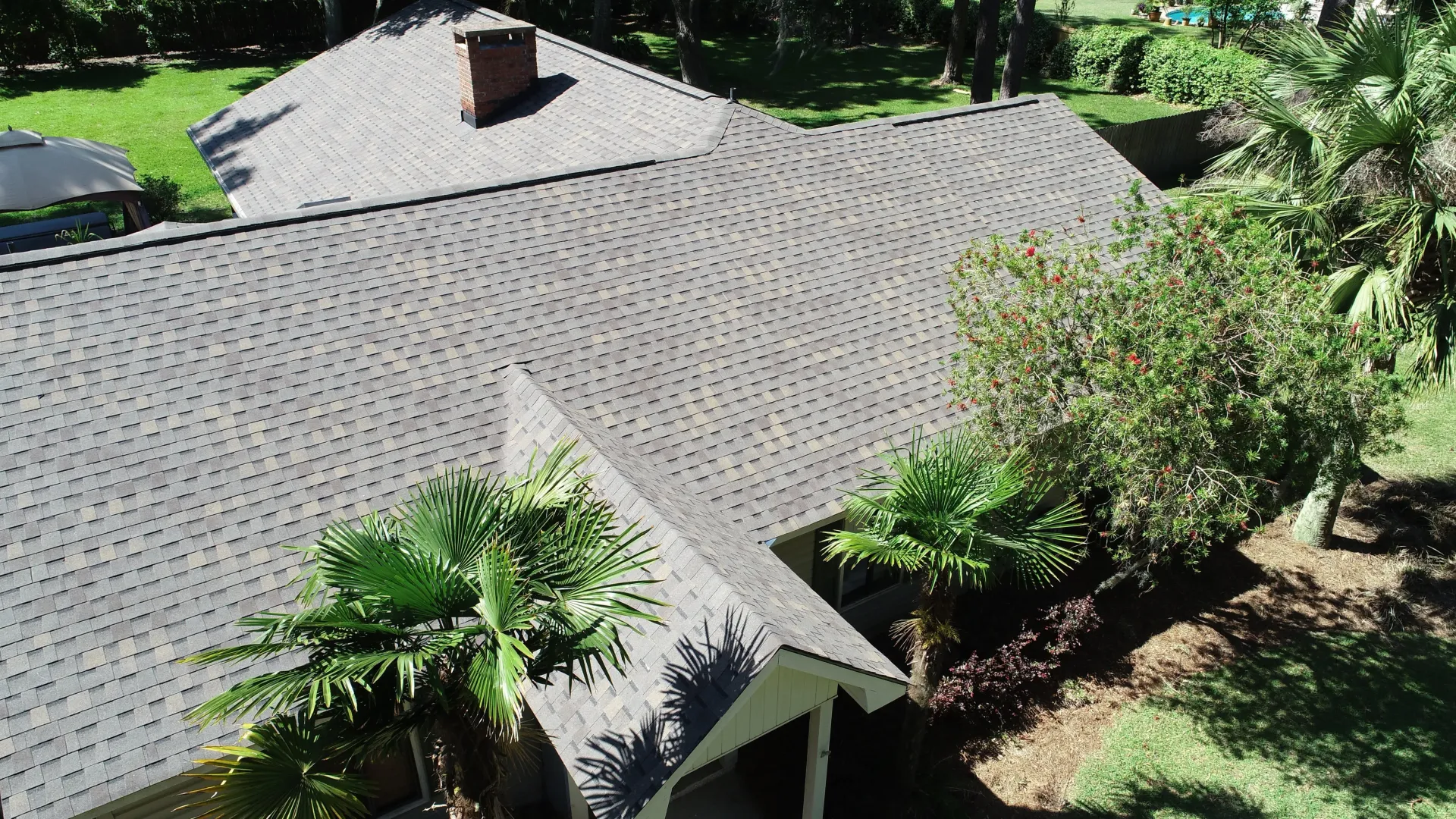 House roof with gray shingles surrounded by lush green trees and shrubs.