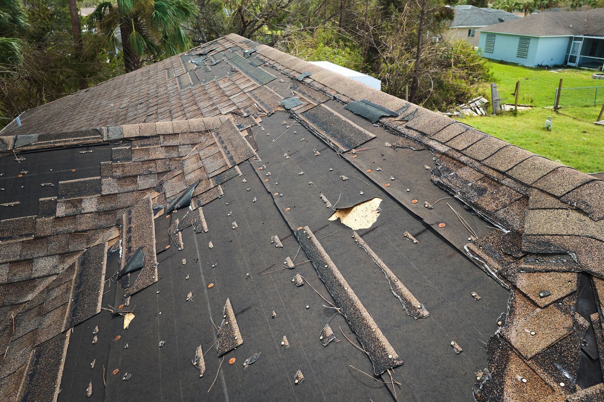 Damaged shingle roof with missing sections and scattered debris on a house near trees and a yard