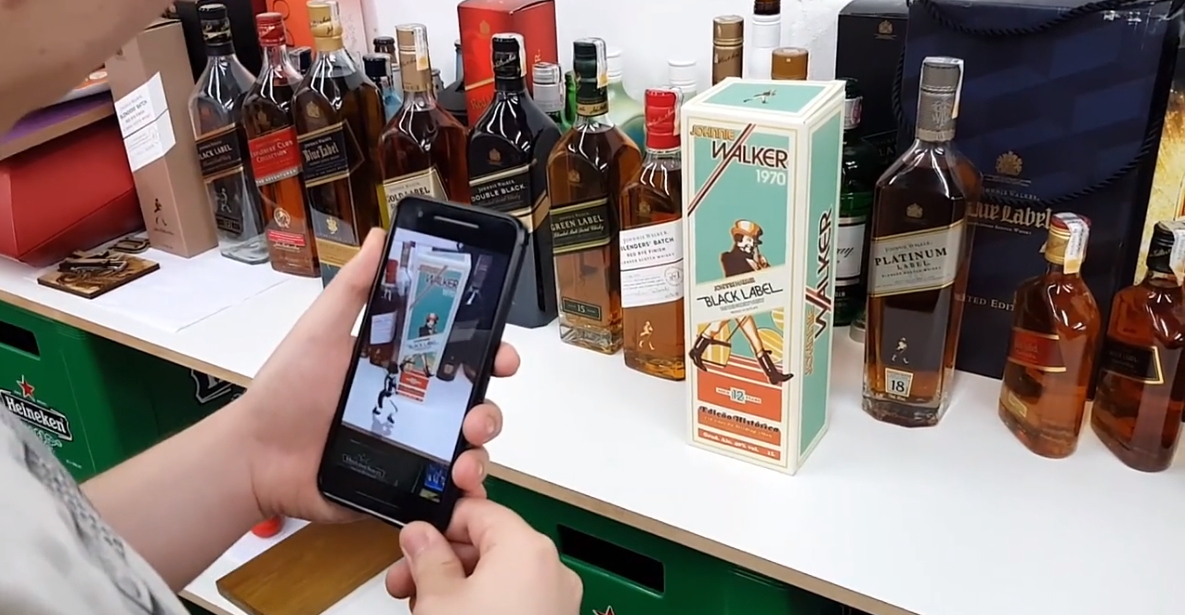 Uma pessoa está segurando um celular em frente a uma mesa cheia de garrafas de álcool.