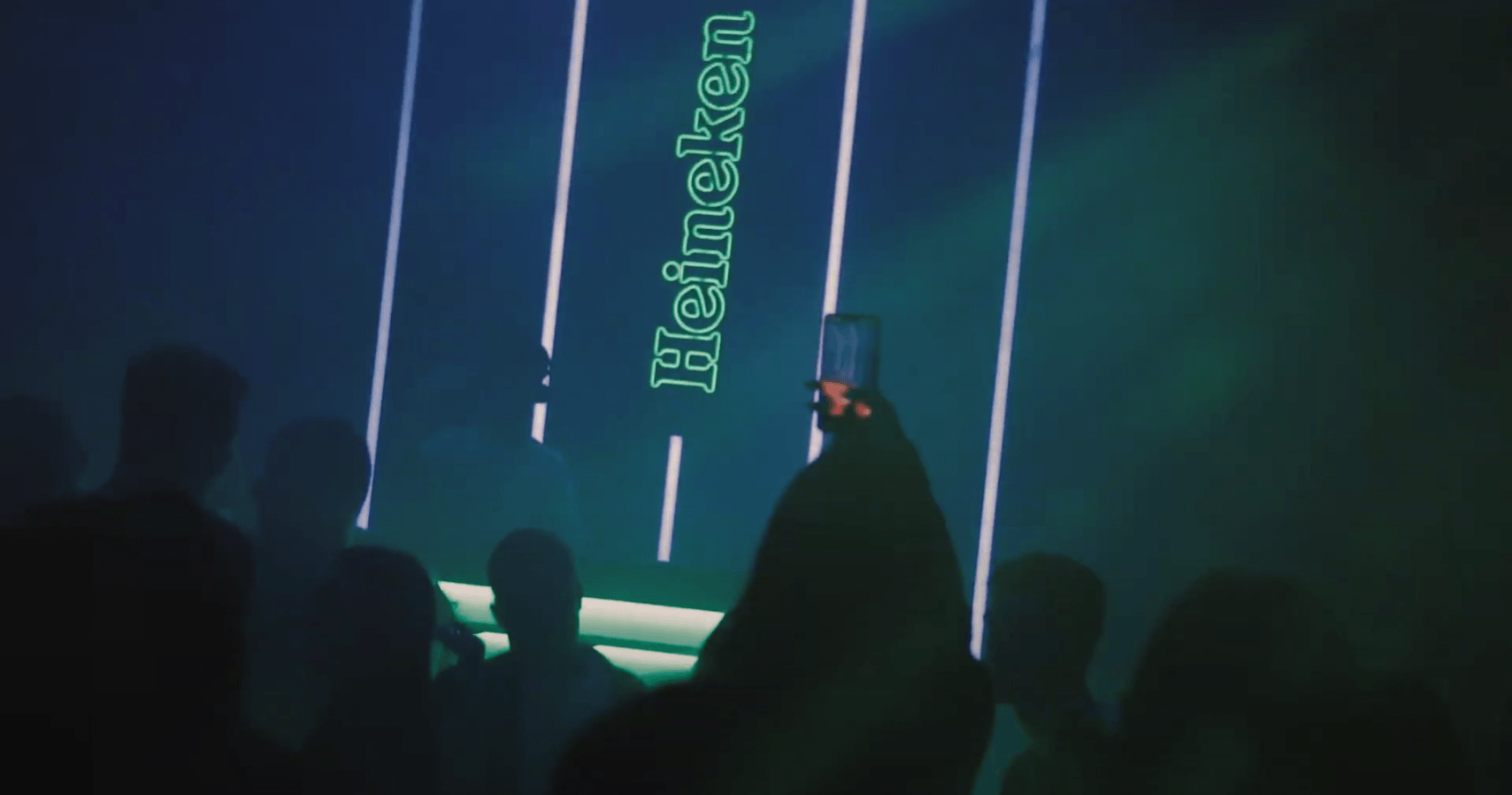 Um grupo de pessoas está em frente a uma placa de neon que diz heineken.