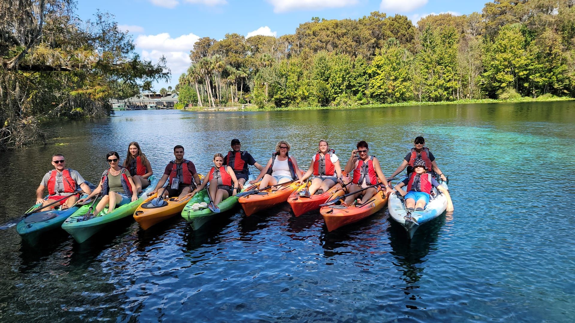 Group kayaking on a sunny river. People wear life vests. Trees line the banks with blue sky.