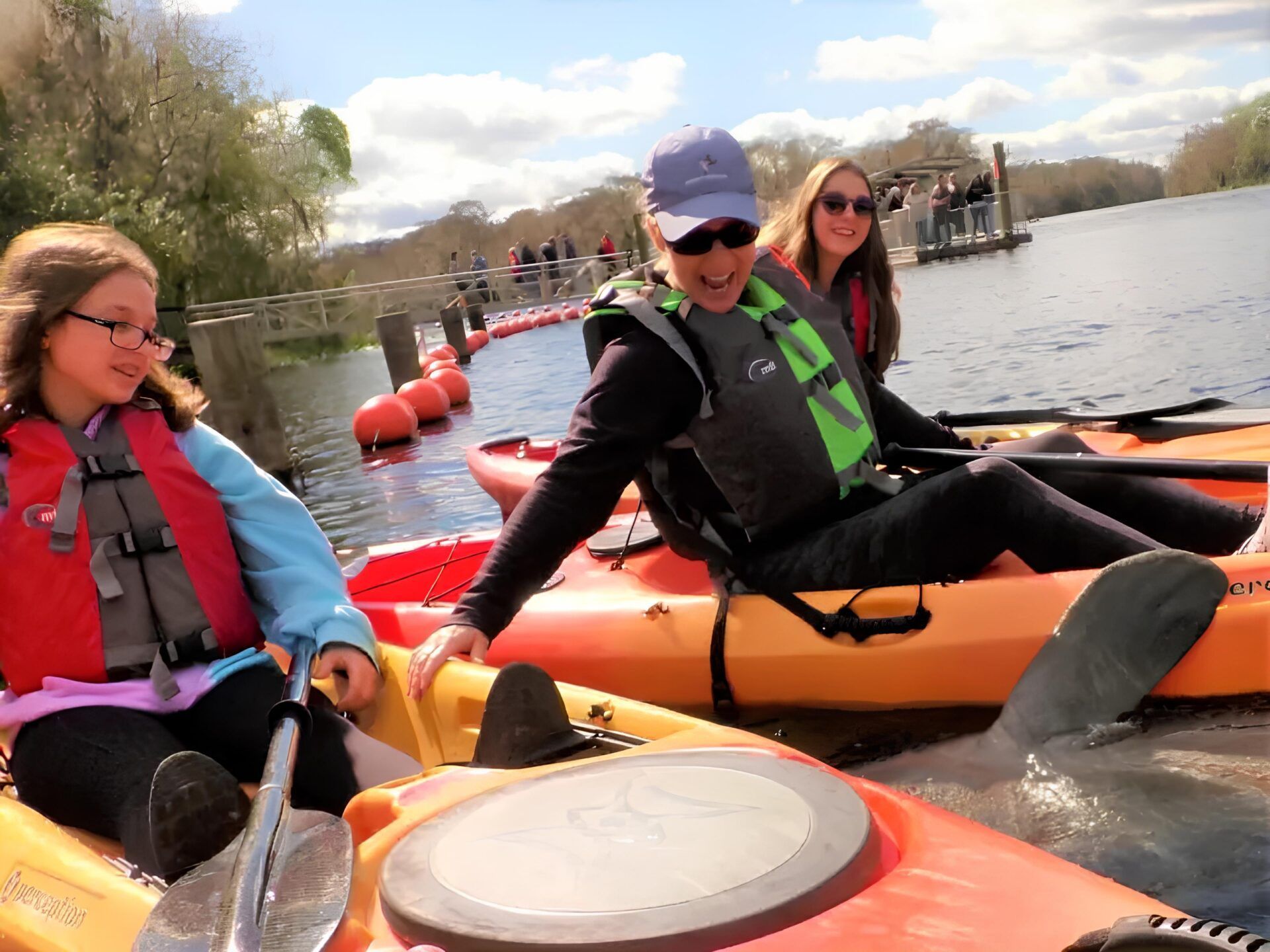 Three people in kayaks near a dock, wearing life vests. Sunny day, smiling.