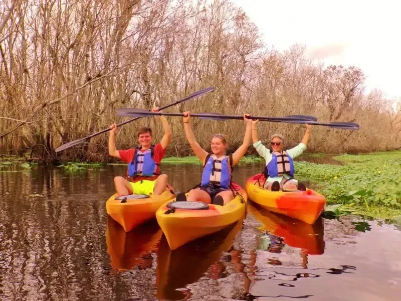 Three people in kayaks raise paddles in a river lined with trees; all wear life vests, smiling.