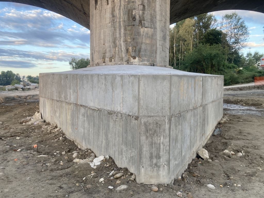 Base di sostegno del ponte in cemento, grigia e usurata, su terreno sterrato.