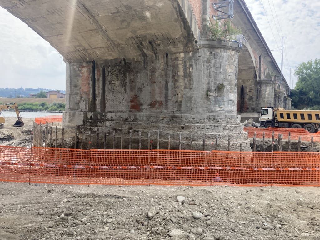 Pilastro del ponte in costruzione, con recinzione di sicurezza arancione. Macchine edili e fiume sullo sfondo.