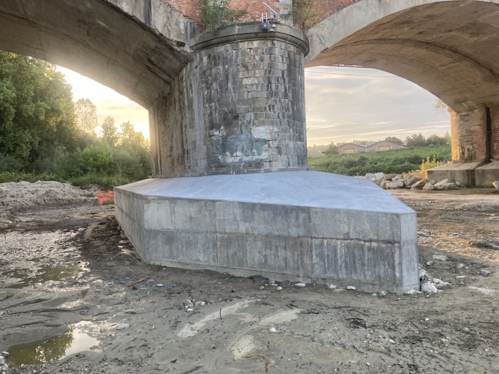 Pilastro del ponte con nuova base in cemento, in un letto di fiume fangoso. Archi in alto, fogliame verde sullo sfondo.