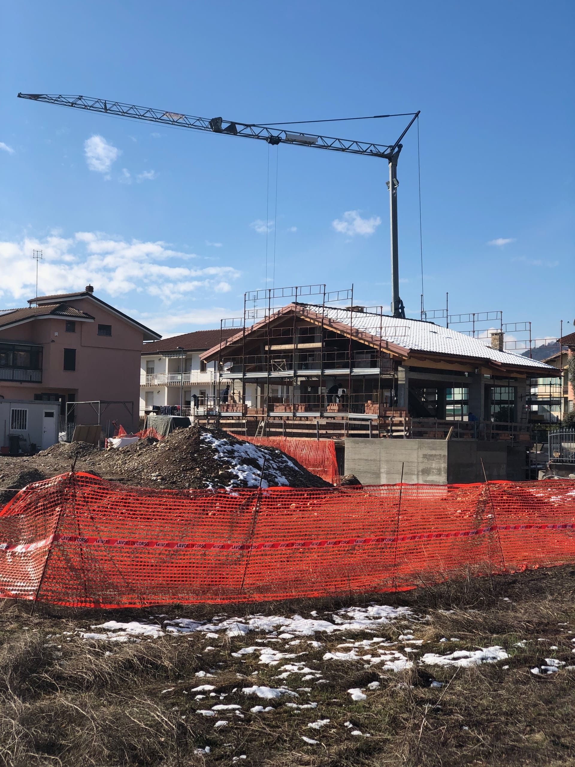 Cantiere con gru e struttura portante dell'edificio; recinzione di sicurezza arancione di fronte.